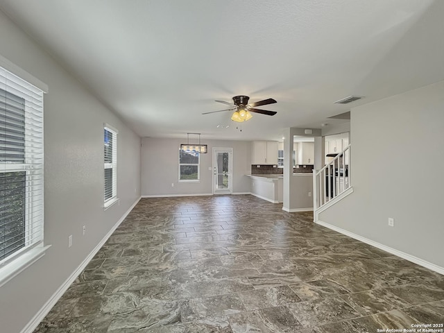 unfurnished living room featuring visible vents, baseboards, stairs, and ceiling fan with notable chandelier