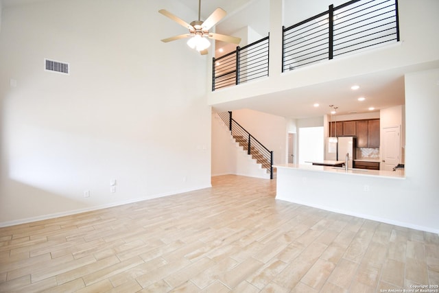 unfurnished living room featuring visible vents, a ceiling fan, a high ceiling, light wood finished floors, and stairs