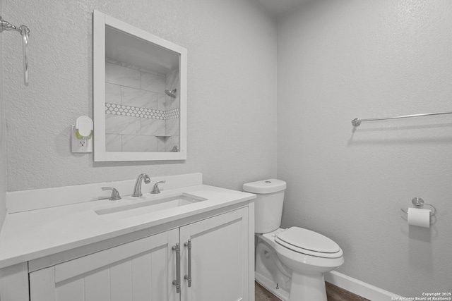 bathroom with toilet, wood finished floors, tiled shower, vanity, and a textured wall