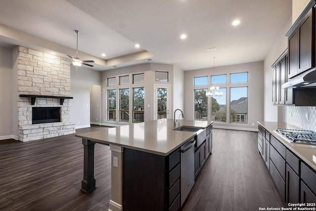 kitchen with dark wood finished floors, an island with sink, a stone fireplace, appliances with stainless steel finishes, and a sink