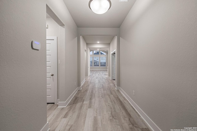 hall featuring a textured wall, baseboards, and light wood finished floors