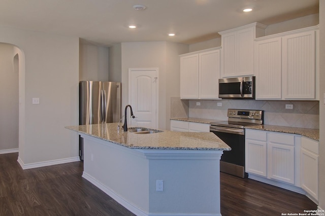 kitchen featuring an island with sink, a sink, stainless steel appliances, arched walkways, and dark wood-style flooring