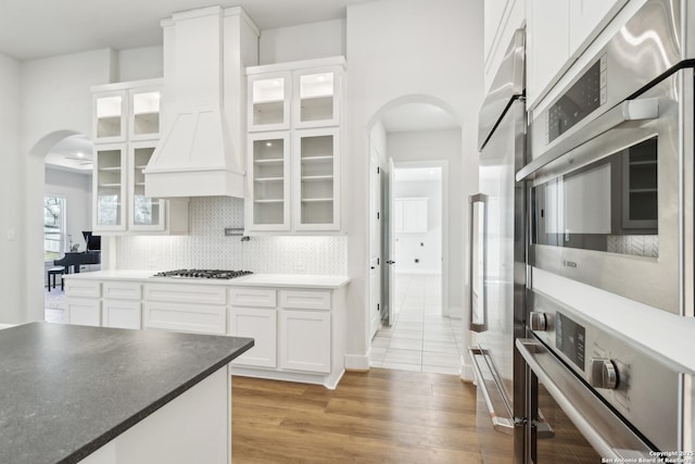 kitchen with custom range hood, backsplash, white cabinetry, arched walkways, and appliances with stainless steel finishes