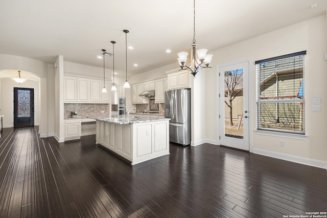 kitchen featuring decorative backsplash, dark wood-style flooring, appliances with stainless steel finishes, arched walkways, and a kitchen island with sink