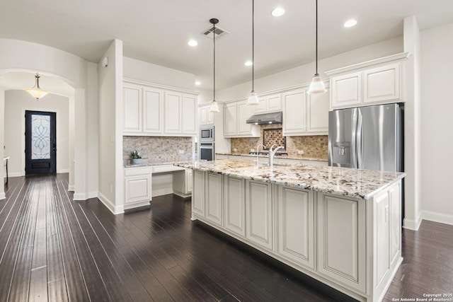 kitchen featuring tasteful backsplash, under cabinet range hood, a center island with sink, appliances with stainless steel finishes, and arched walkways