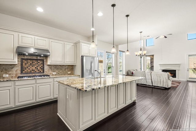 kitchen featuring dark wood-type flooring, under cabinet range hood, a warm lit fireplace, appliances with stainless steel finishes, and decorative backsplash