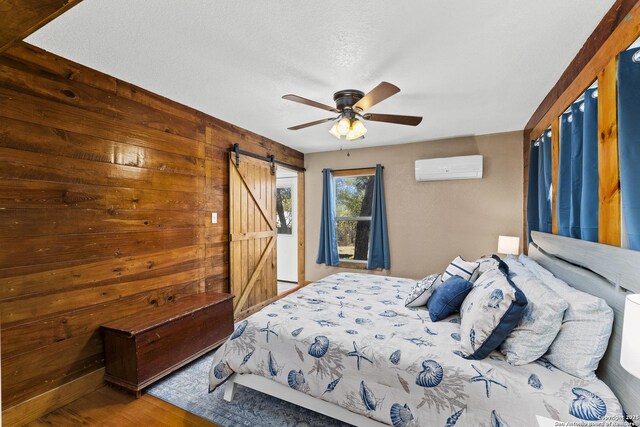 bedroom with an AC wall unit, wood finished floors, a barn door, wood walls, and ceiling fan