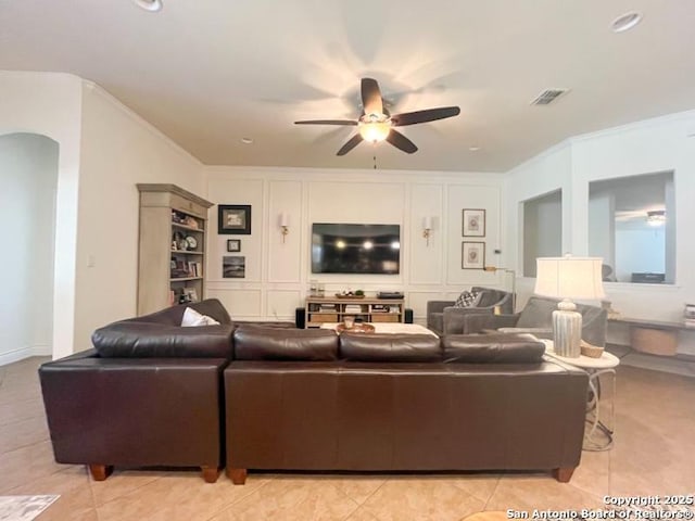 living area featuring light tile patterned floors, a ceiling fan, arched walkways, ornamental molding, and a decorative wall