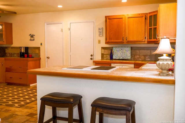 kitchen with a kitchen bar, recessed lighting, brown cabinetry, glass insert cabinets, and tile counters