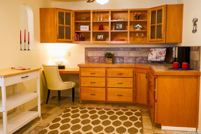 kitchen featuring brown cabinetry, built in study area, open shelves, tile counters, and glass insert cabinets