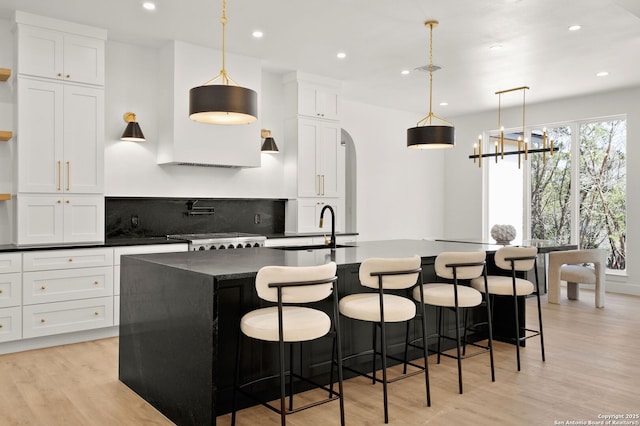 kitchen featuring dark countertops, a sink, light wood-type flooring, arched walkways, and a kitchen island with sink