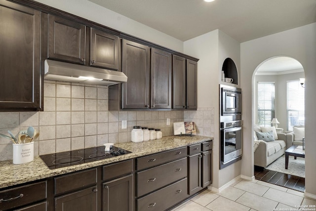 kitchen featuring dark brown cabinets, under cabinet range hood, light stone counters, light tile patterned flooring, and stainless steel appliances