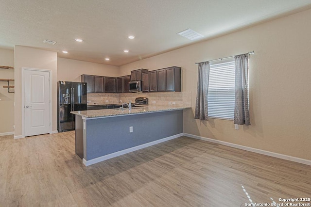 kitchen with stainless steel microwave, black fridge with ice dispenser, light wood-style floors, decorative backsplash, and light stone countertops