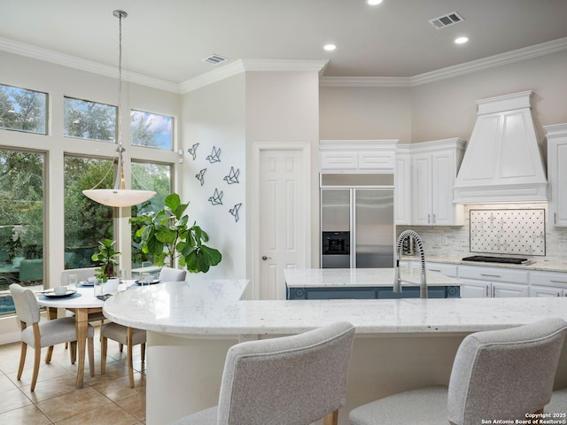 kitchen featuring visible vents, premium range hood, built in refrigerator, an island with sink, and decorative backsplash