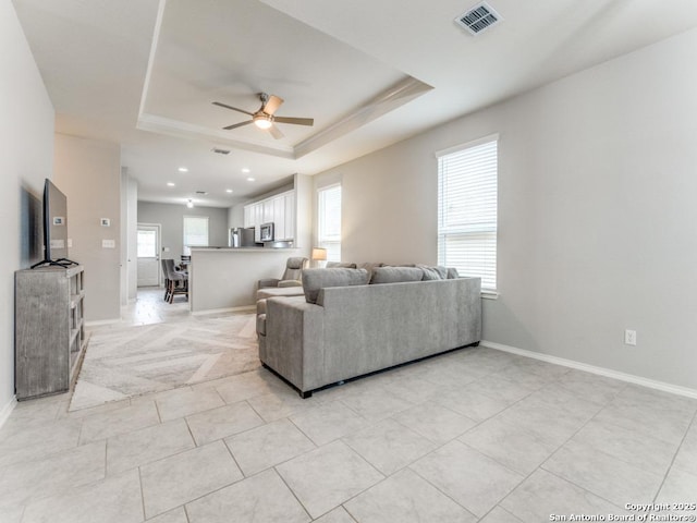 living area with a tray ceiling, baseboards, visible vents, and ceiling fan