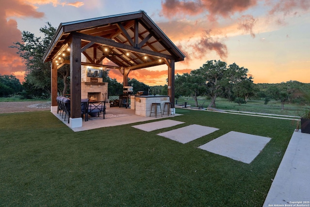 yard at dusk with outdoor dry bar, a gazebo, an outdoor stone fireplace, area for grilling, and a patio