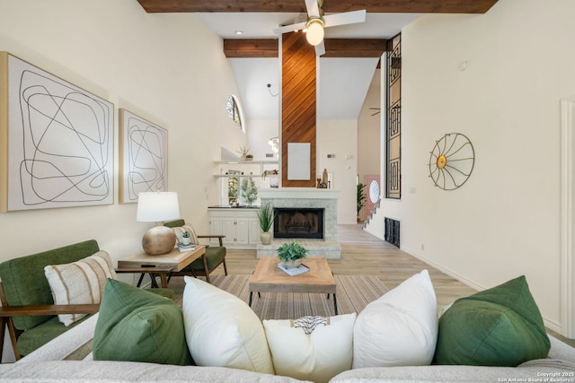 living area with beam ceiling, high vaulted ceiling, a fireplace with raised hearth, a ceiling fan, and wood finished floors