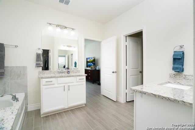 full bath featuring wood finished floors, visible vents, baseboards, two vanities, and a garden tub