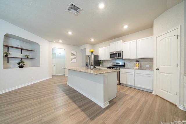 kitchen featuring visible vents, light wood-style flooring, an island with sink, arched walkways, and appliances with stainless steel finishes