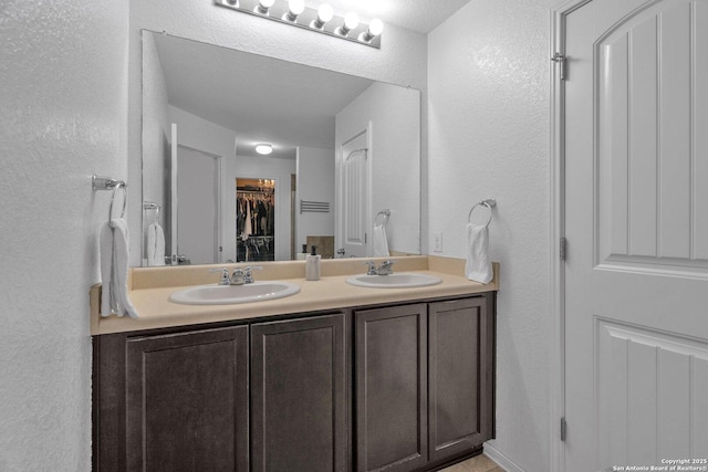 full bath with double vanity, a walk in closet, a textured wall, and a sink