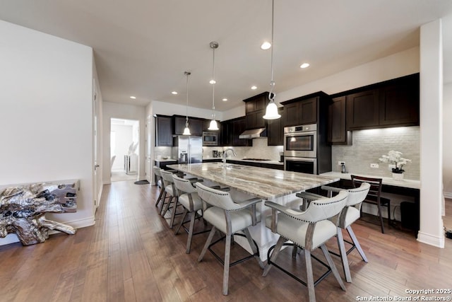 kitchen featuring a breakfast bar, under cabinet range hood, backsplash, appliances with stainless steel finishes, and light wood finished floors