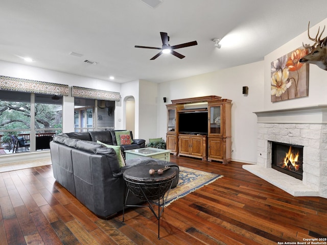 living room featuring visible vents, a ceiling fan, dark wood-style floors, arched walkways, and a stone fireplace