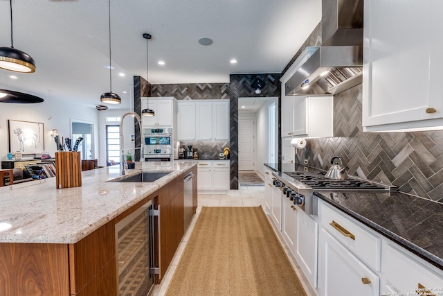 kitchen with backsplash, wall chimney range hood, light stone countertops, beverage cooler, and a large island with sink