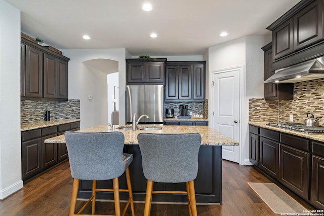 kitchen featuring a kitchen island with sink, arched walkways, dark wood-style floors, stainless steel appliances, and a sink