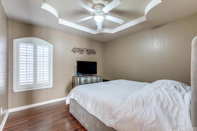 bedroom featuring baseboards, a tray ceiling, dark wood-style flooring, ceiling fan, and a textured wall