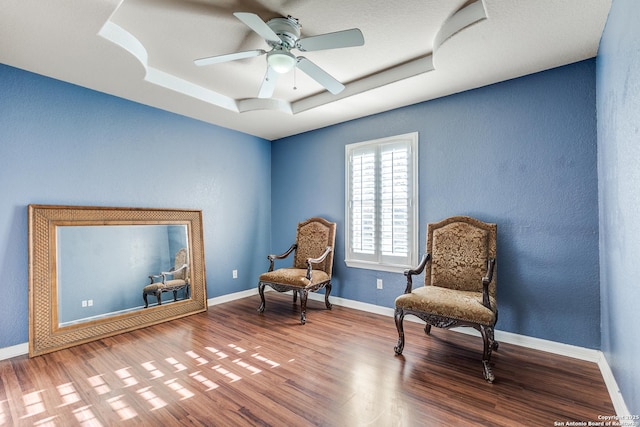 sitting room with baseboards, wood finished floors, a textured wall, a raised ceiling, and a ceiling fan