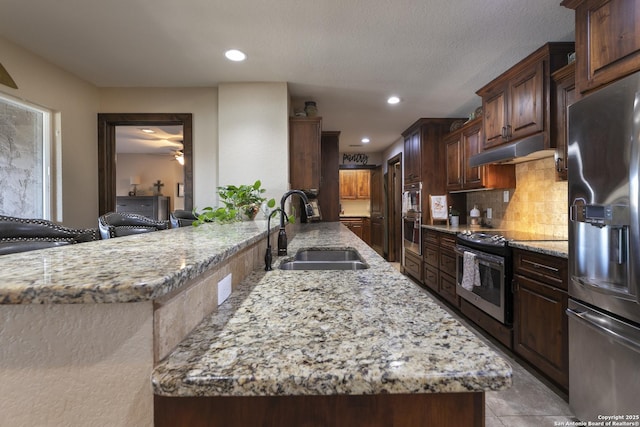 kitchen with tasteful backsplash, light stone countertops, under cabinet range hood, stainless steel appliances, and a sink