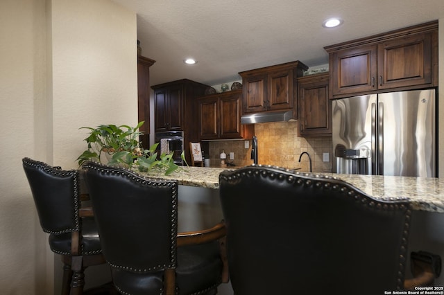 kitchen with under cabinet range hood, backsplash, dark brown cabinetry, stainless steel fridge with ice dispenser, and light stone countertops