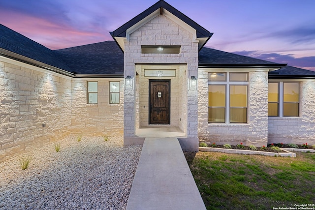 exterior entry at dusk featuring stone siding and a shingled roof