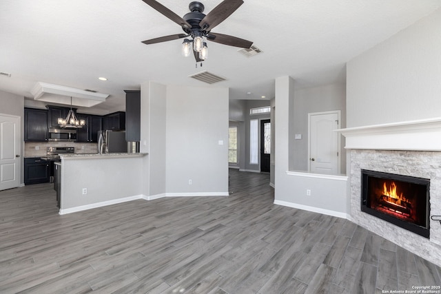 unfurnished living room with visible vents, baseboards, a stone fireplace, ceiling fan with notable chandelier, and light wood-style floors
