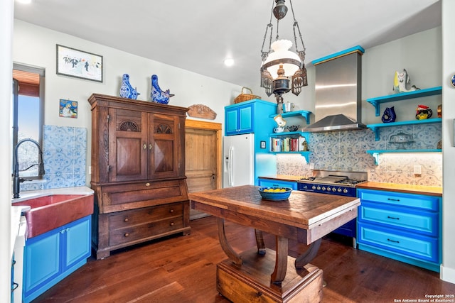 kitchen with blue cabinets, white fridge with ice dispenser, wall chimney exhaust hood, decorative backsplash, and dark wood-style flooring