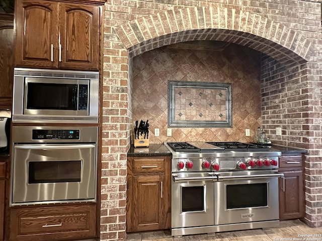 kitchen featuring tasteful backsplash, brick wall, appliances with stainless steel finishes, and dark stone counters