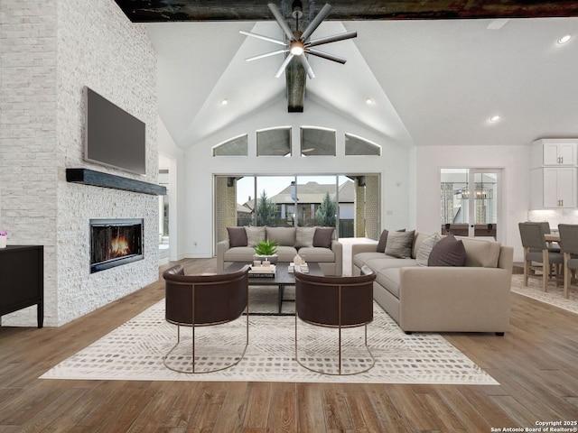 living area featuring beam ceiling, high vaulted ceiling, wood finished floors, a stone fireplace, and ceiling fan