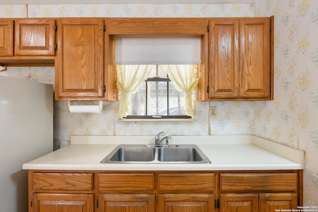 kitchen featuring wallpapered walls, light countertops, brown cabinetry, and a sink