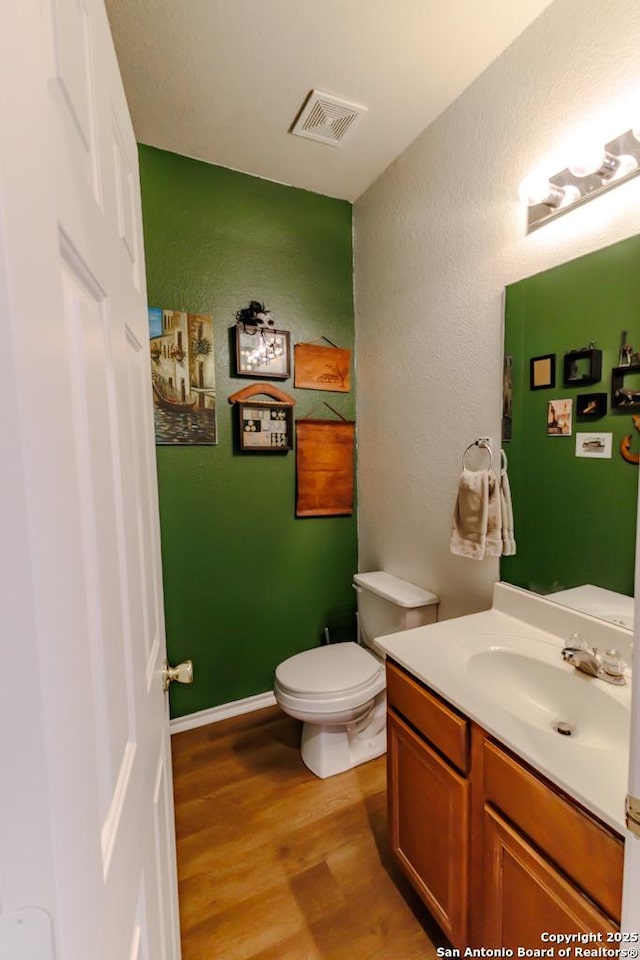 half bathroom with vanity, wood finished floors, visible vents, toilet, and a textured wall