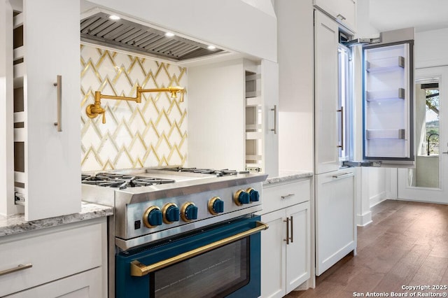 kitchen with light stone countertops, wood finished floors, custom range hood, white cabinets, and high end stainless steel range oven