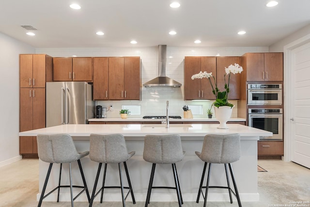 kitchen with a kitchen island with sink, decorative backsplash, appliances with stainless steel finishes, wall chimney exhaust hood, and brown cabinets