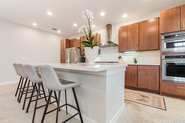 kitchen with backsplash, wall chimney range hood, double oven, a kitchen bar, and a sink