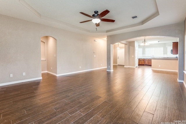 unfurnished living room with a raised ceiling, ceiling fan with notable chandelier, visible vents, and dark wood-type flooring