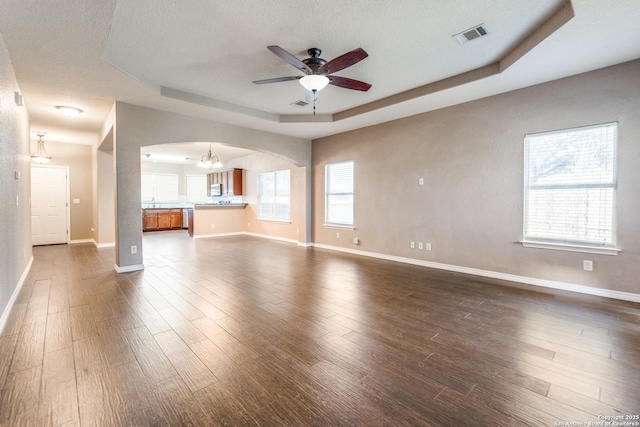 unfurnished living room with arched walkways, ceiling fan with notable chandelier, a tray ceiling, and dark wood-style flooring