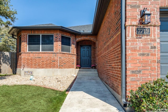 view of exterior entry with brick siding, a shingled roof, and fence