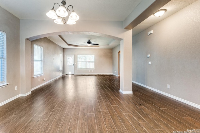 unfurnished living room featuring wood finished floors, baseboards, a tray ceiling, arched walkways, and ceiling fan with notable chandelier