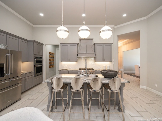 kitchen featuring tasteful backsplash, premium range hood, a center island with sink, gray cabinets, and stainless steel appliances