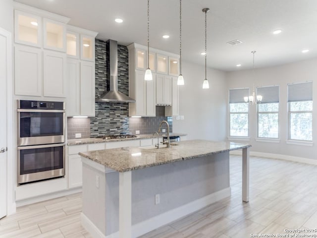 kitchen with backsplash, a center island with sink, white cabinets, stainless steel appliances, and wall chimney exhaust hood