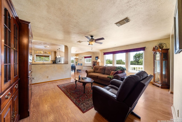 living room with visible vents, light wood-style flooring, ceiling fan with notable chandelier, arched walkways, and a textured ceiling