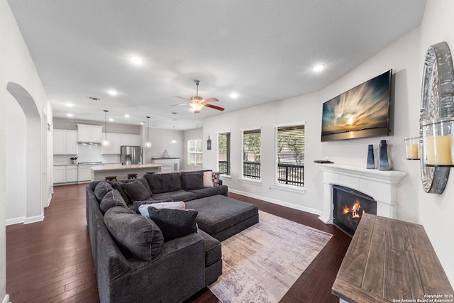 living area with a glass covered fireplace, baseboards, dark wood-style floors, and a ceiling fan